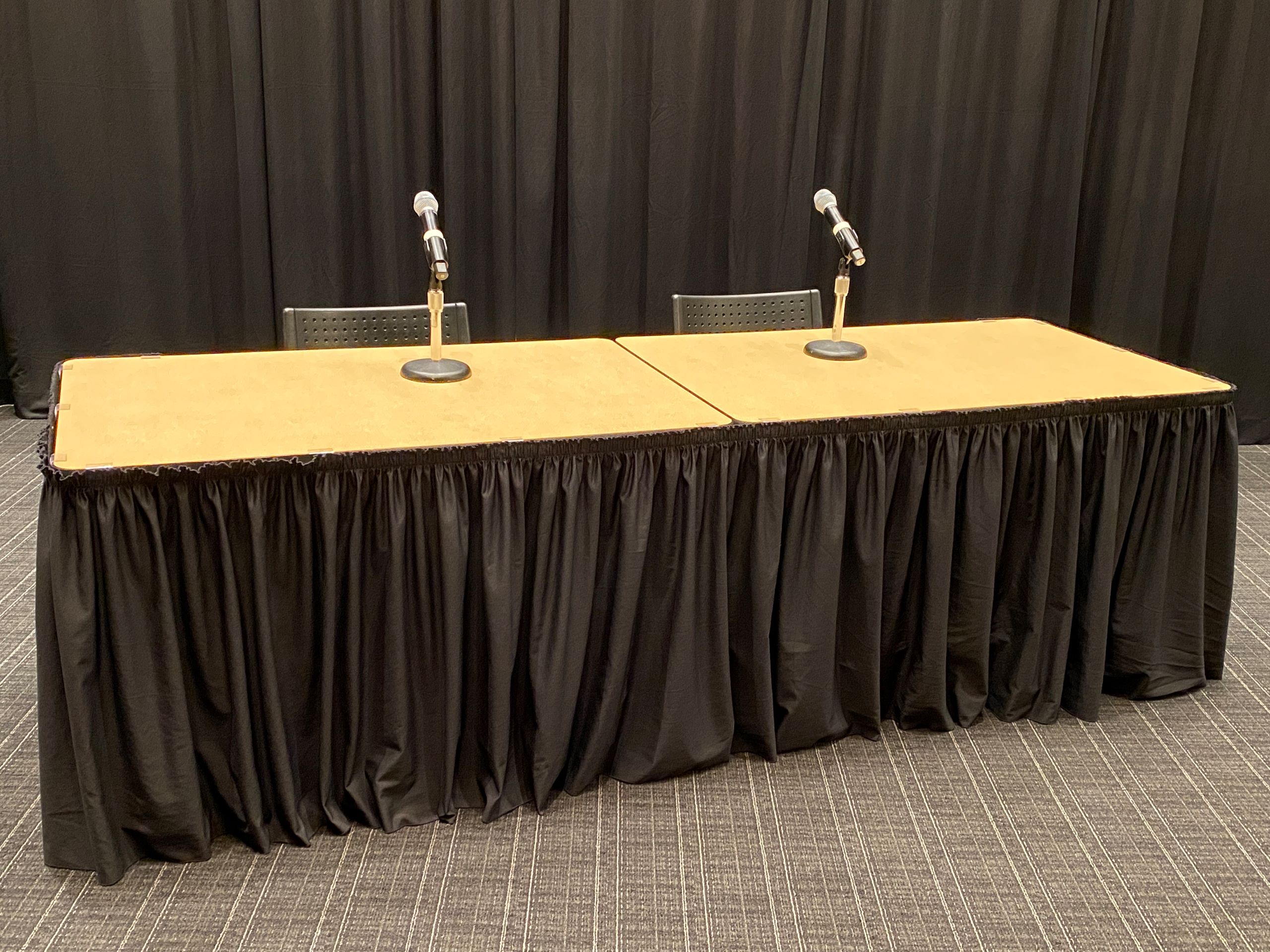 A wooden-top folding table with black table skirting around the edges and two microphones on mic stands to simulate a panel setup.