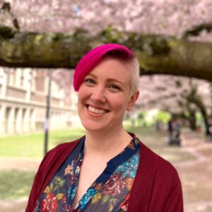 Portrait of Jessilee Marander, Event Services Program Coordinator, beaming with bright pink hair in front of cherry blossom trees.